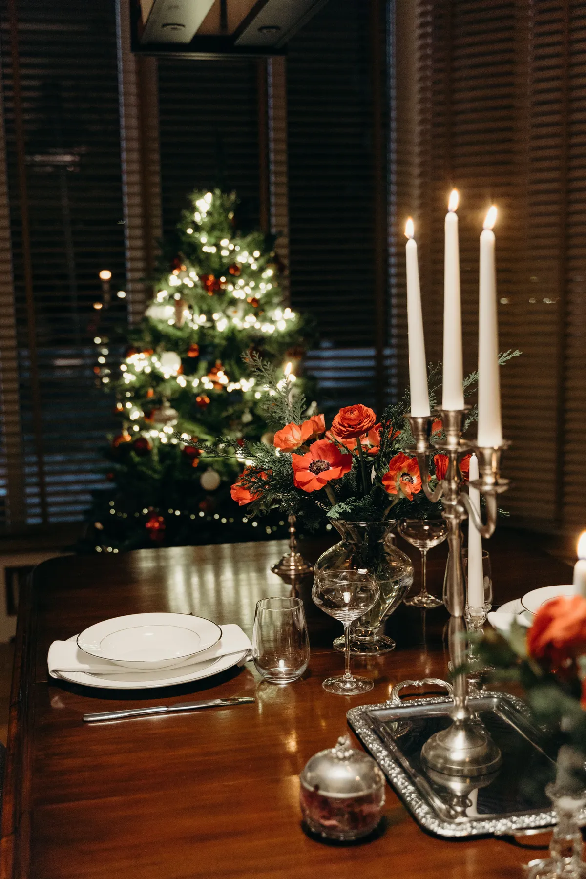 A warm, decorative Christmas table setting with candlelight, flowers, and a decorated tree.