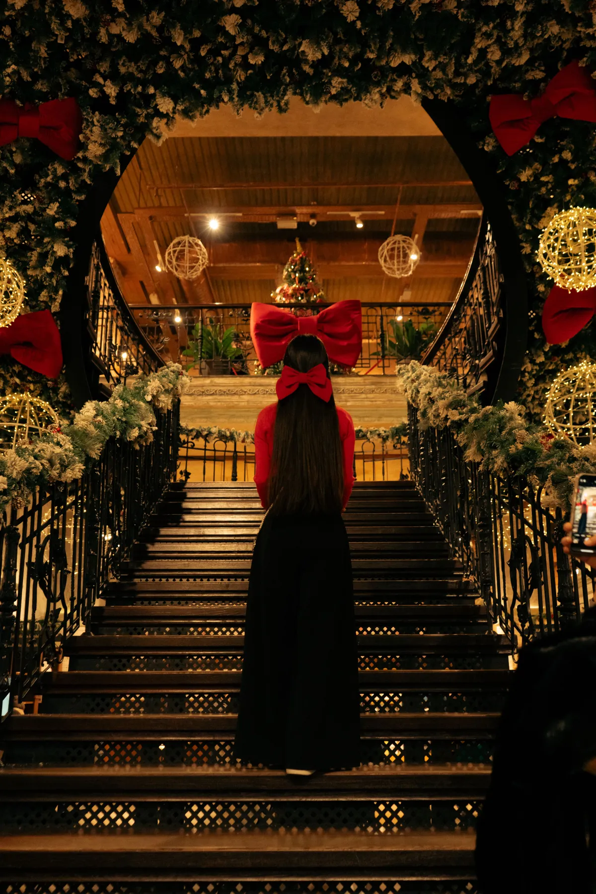 A woman in a festive outfit stands on beautifully decorated stairs with Christmas lights and bows.