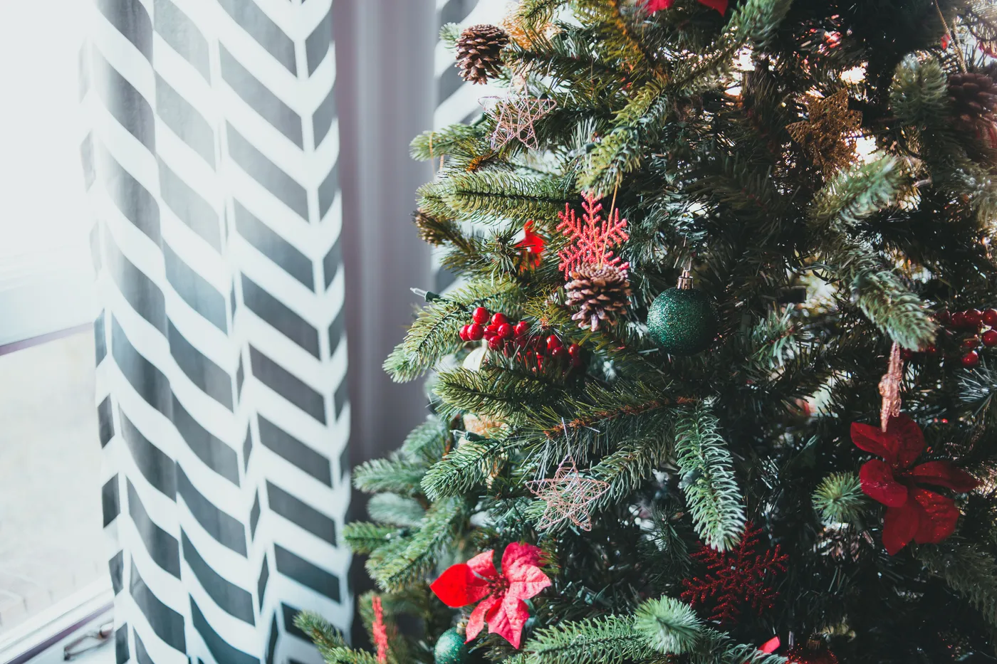 Christmas tree decorated with toys standing near window with patterned curtain on daylight in winter