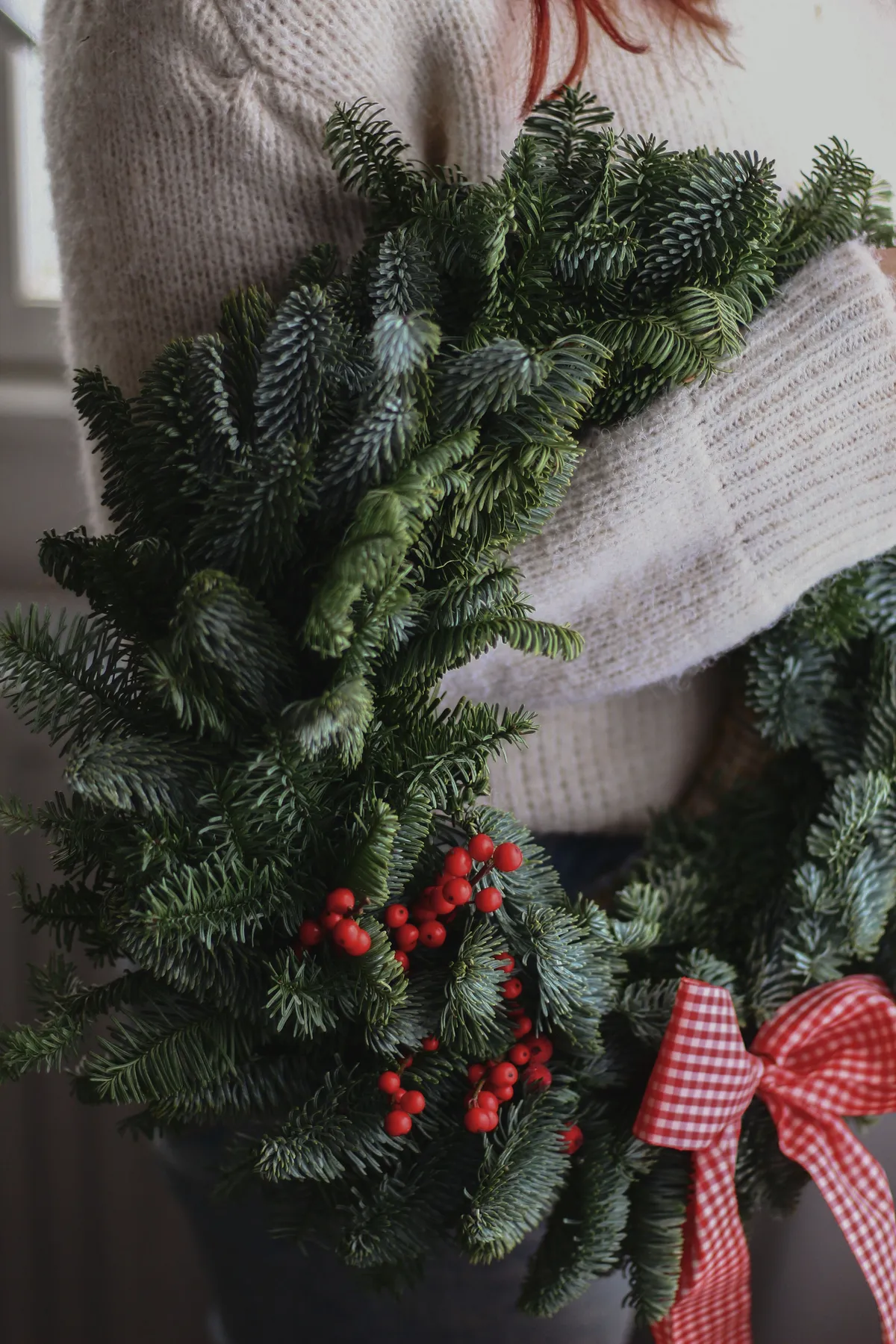 Close-up of a Christmas wreath with red berries and a gingham bow held by a person in a knitted sweater.
