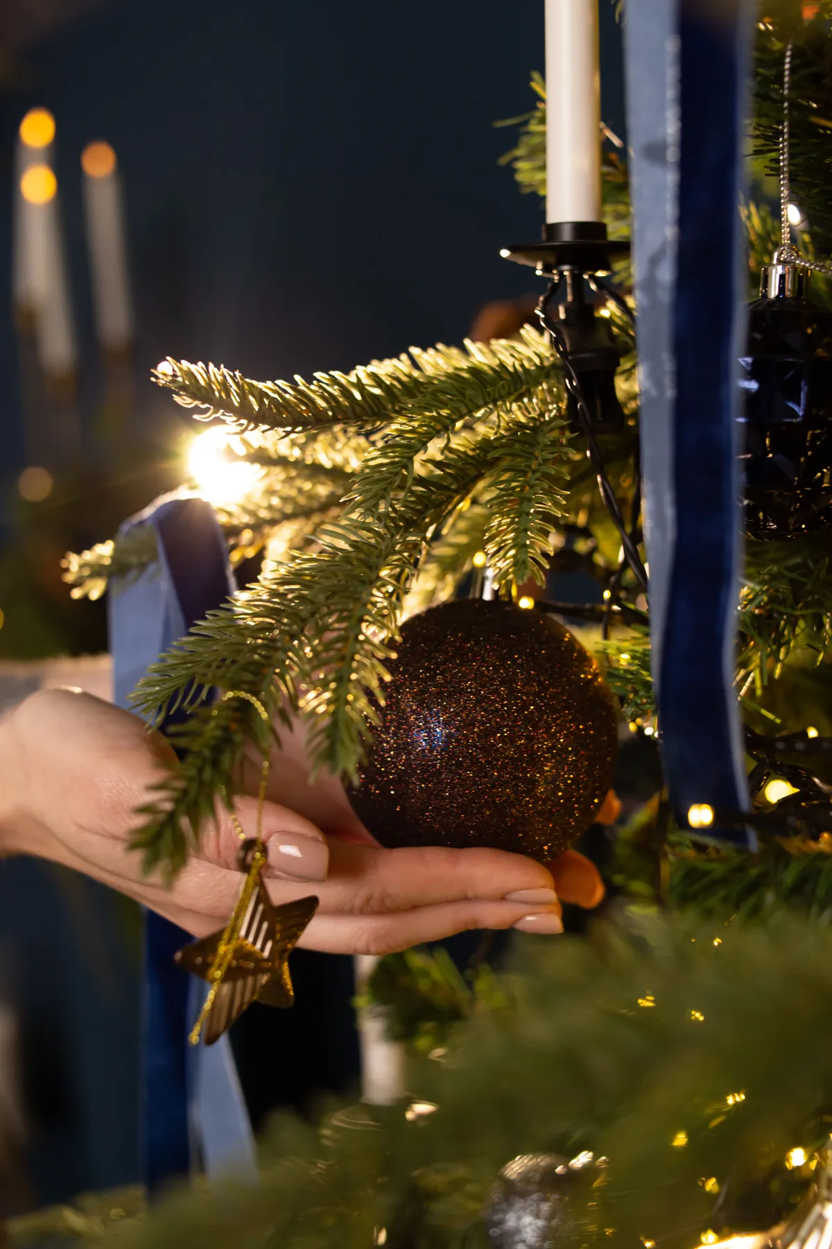 Close-up of a hand placing a glittery bauble on a Christmas tree branch with candles.