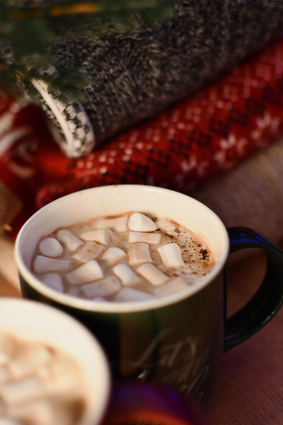 Cozy cup of hot chocolate topped with marshmallows next to festive blankets.