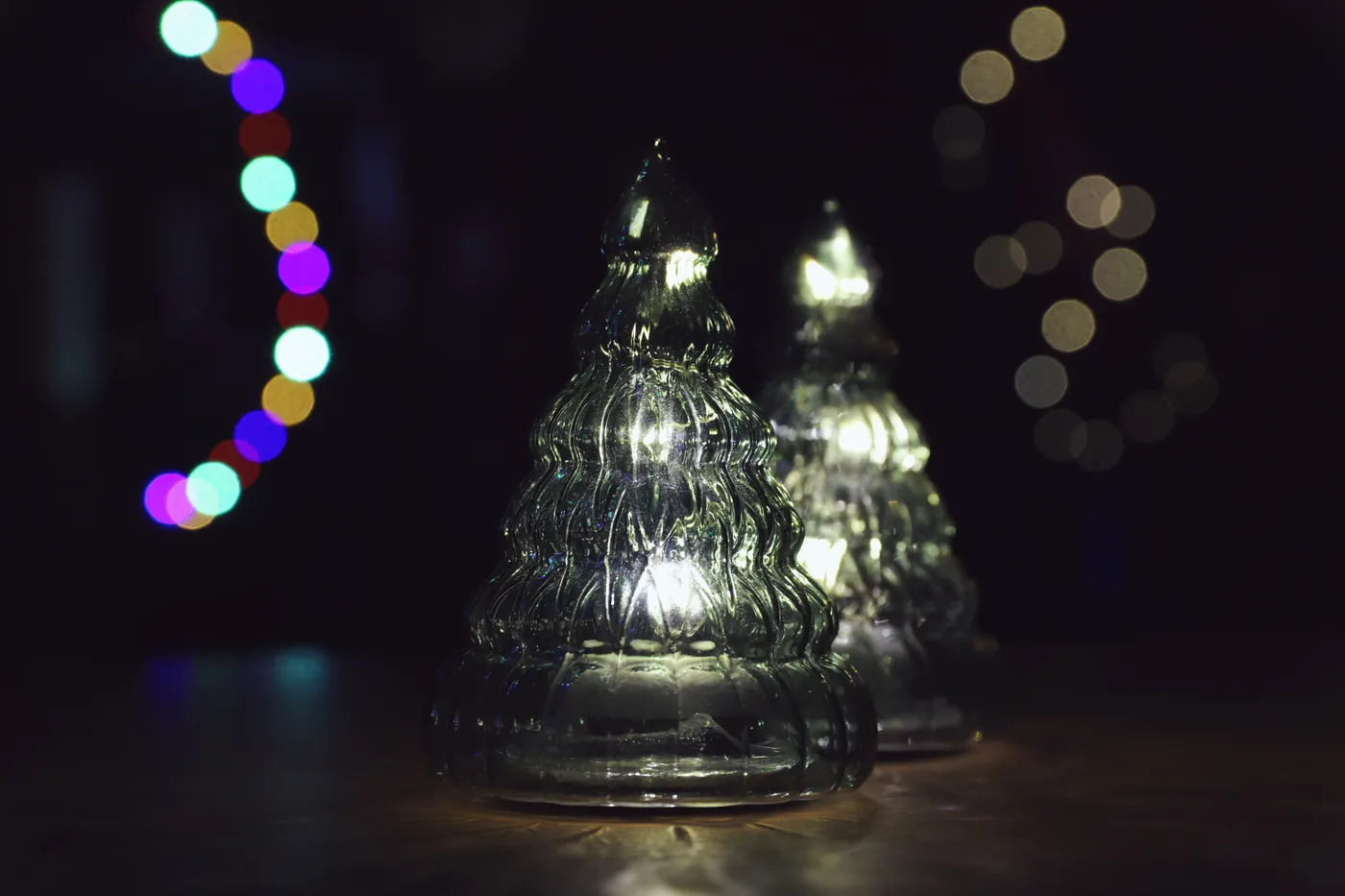Illuminated glass Christmas trees with colorful bokeh lights against a dark background.
