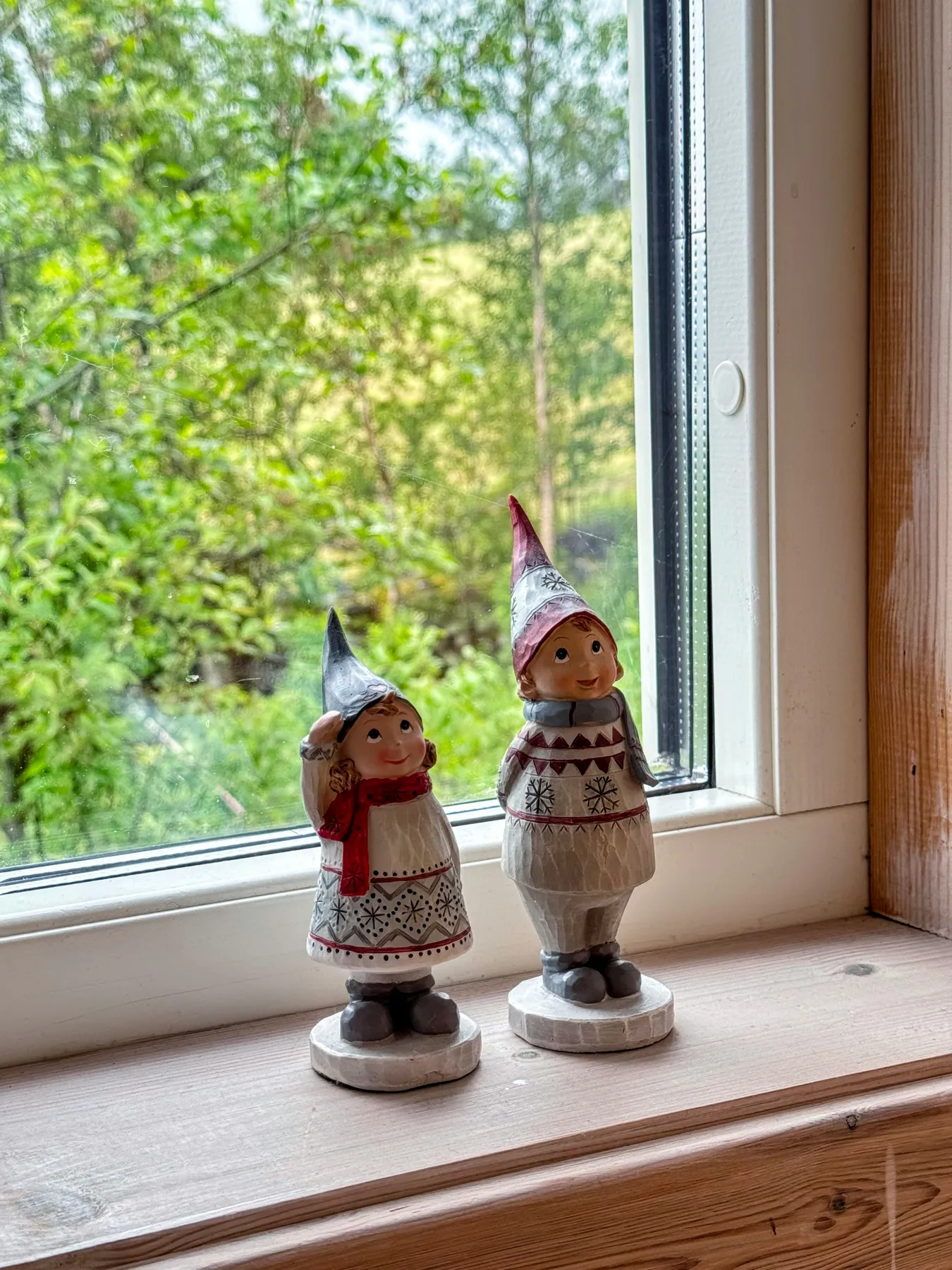 Two small gnome figurines in winter clothing on a wooden windowsill before green trees